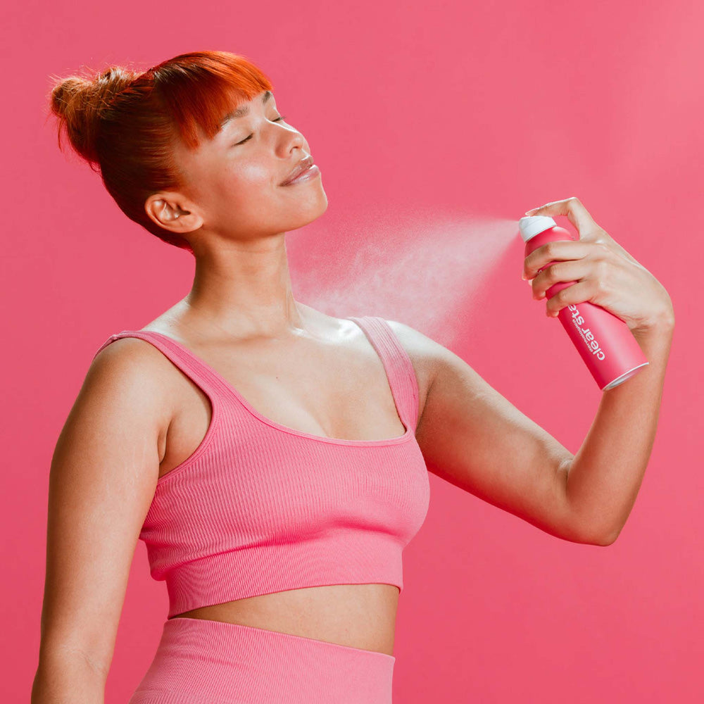 Woman in pink sports bra and shorts applying a spray product against a pink background